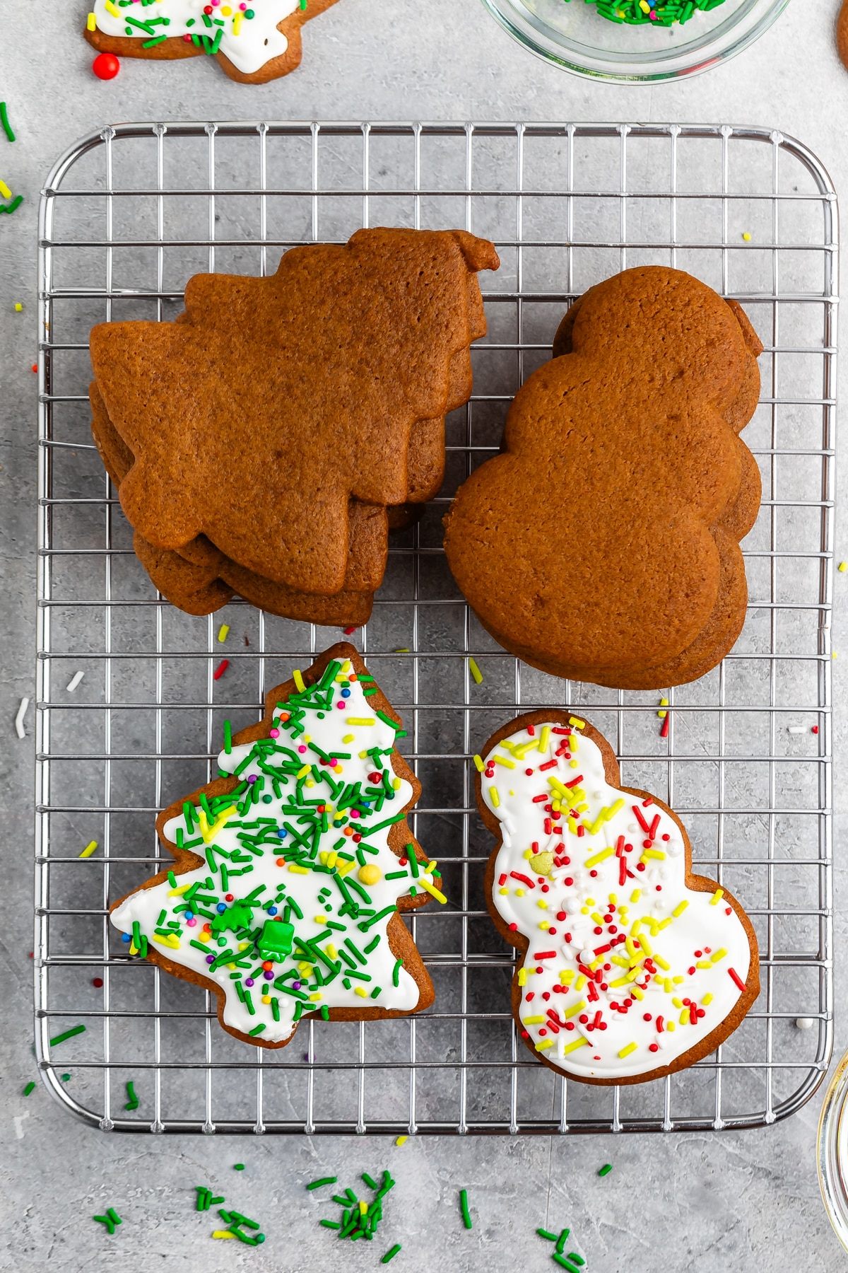 A cooling rack with gingerbread cookies shaped like Christmas trees and snowmen. Some cookies are plain, while others are decorated with white icing and colorful sprinkles. Scattered sprinkles and crumbs surround the rack.