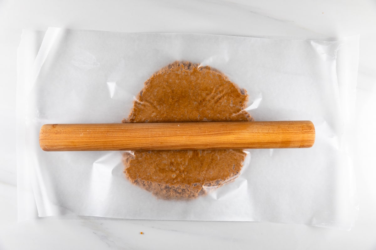 A wooden rolling pin flattening gingerbread cookie dough between two sheets of parchment paper on a white marble surface.