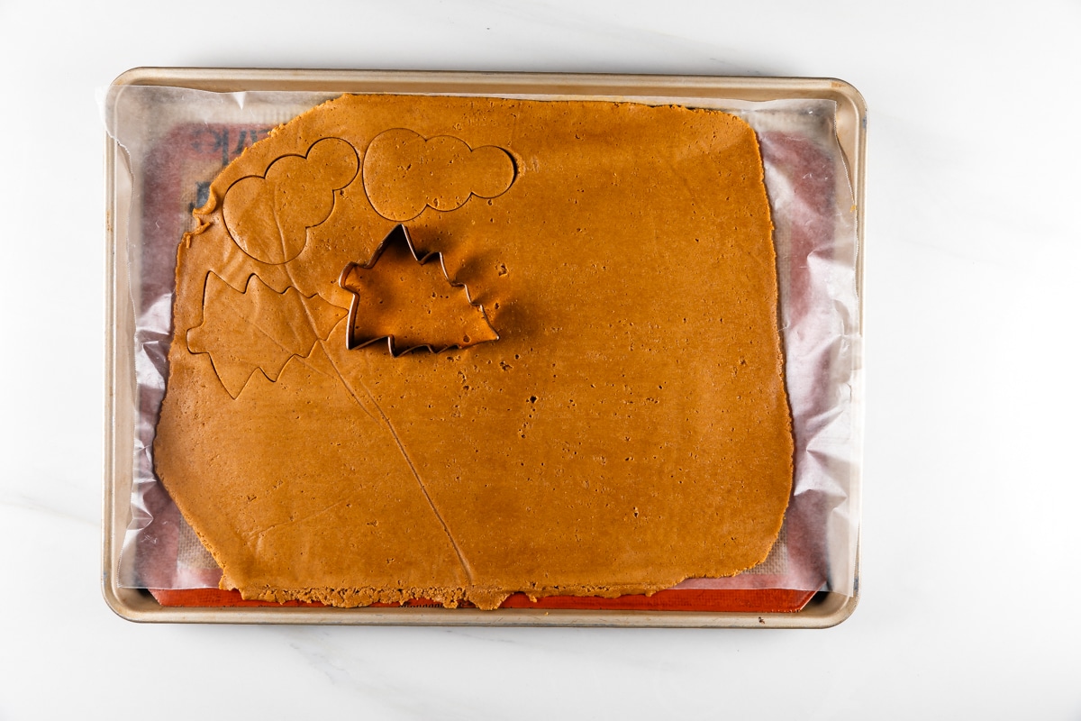 A baking sheet with rolled-out gingerbread dough on parchment paper. Three cookie cutter shapes—a maple leaf, an evergreen tree, and a cloud—are pressed into the dough, with one tree shape cut out.