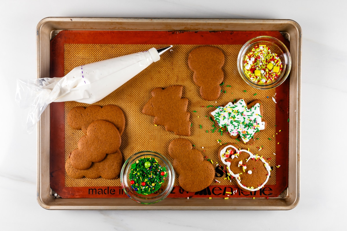 A baking tray with gingerbread cookies shaped like snowmen and trees. Theres a piping bag with white icing and bowls of colorful sprinkles. One cookie is partially decorated with icing and sprinkles.