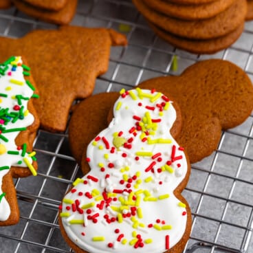 Snowman-shaped gingerbread cookie with white icing and red, yellow, and green sprinkles on a cooling rack. More cookies are visible in the background.