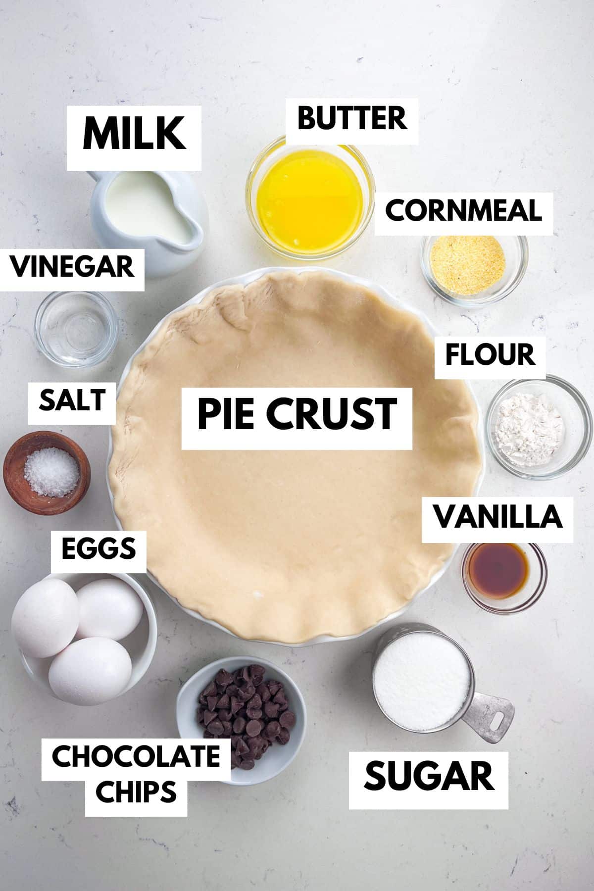 Top view of various pie ingredients labeled around an empty pie crust: milk, butter, cornmeal, vinegar, salt, flour, vanilla, eggs, chocolate chips, and sugar.