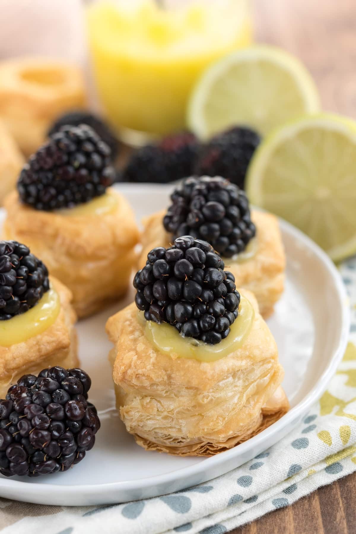 Close-up of puff pastry tarts topped with lemon curd and fresh blackberries on a white plate. In the background, sliced limes and more blackberries are visible, along with a glass of yellow juice. The setting is on a light patterned cloth.