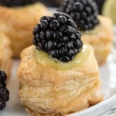 A close-up of a pastry topped with custard and a cluster of fresh blackberries, set on a white plate. The flaky layers of the pastry are visible, and there are more pastries and a slice of lime blurred in the background.