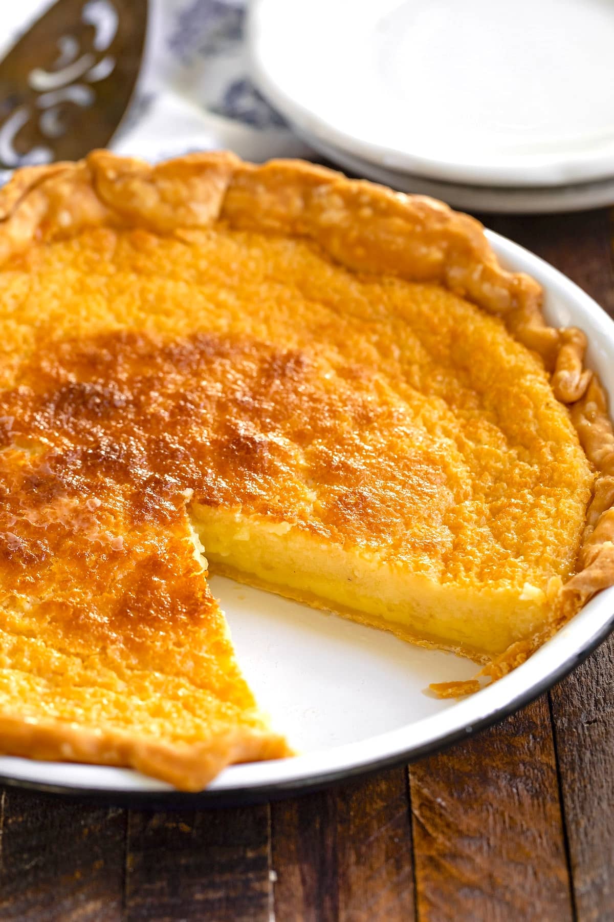 A golden-brown custard pie with a slice missing is displayed on a white dish. The pie has a flaky crust and a smooth, creamy filling. It rests on a wooden table with a serving spatula and a small plate in the background.