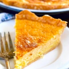 A slice of chess pie with a golden brown crust is served on a white plate next to a fork. The pie has a rich, custard-like texture and is in focus, with the rest of the pie visible in the background.