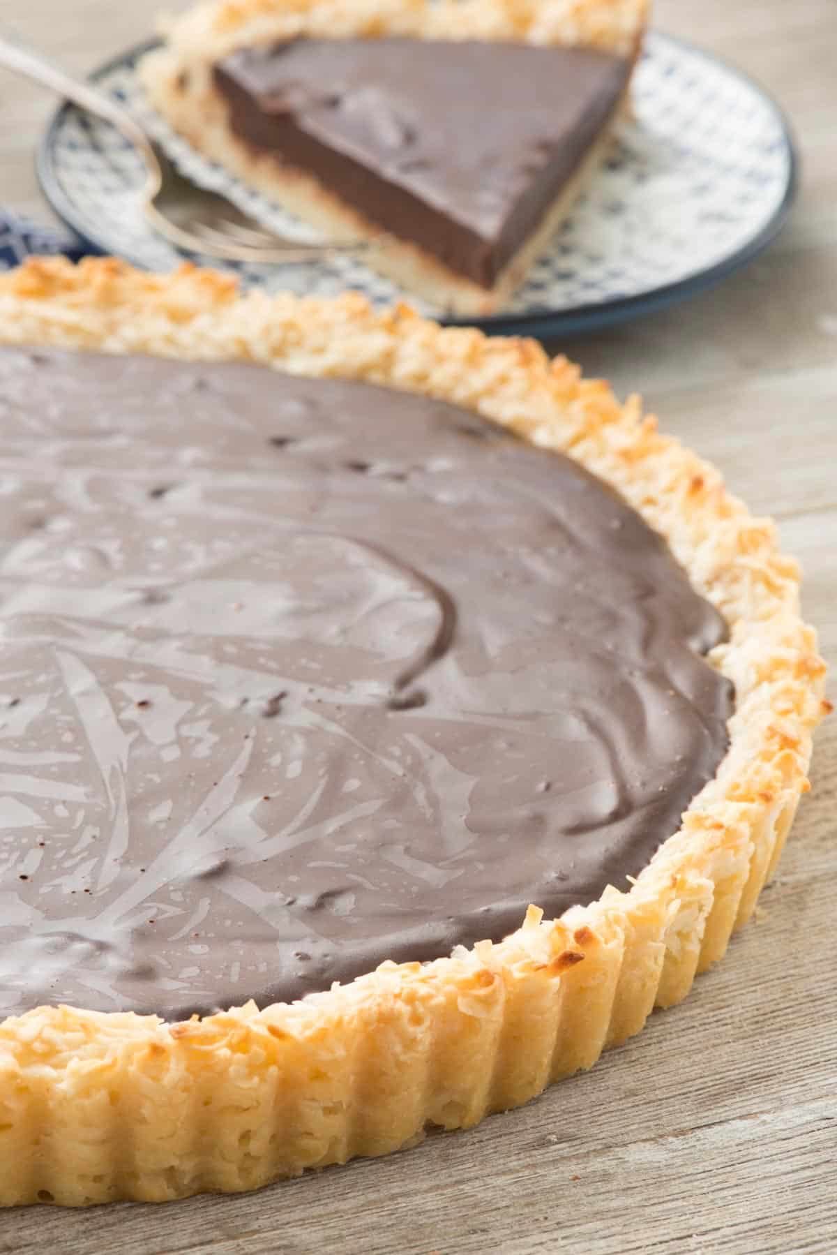 A chocolate tart with a shiny, smooth filling sits in a fluted crust. A slice is served on a patterned plate in the background, with a fork beside it. The wooden surface adds a rustic touch to the setting.