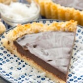 A slice of chocolate tart with a coconut crust sits on a white plate with blue dots. Shredded coconut is scattered around the plate. In the background, more tart slices and a bowl of shredded coconut are visible.