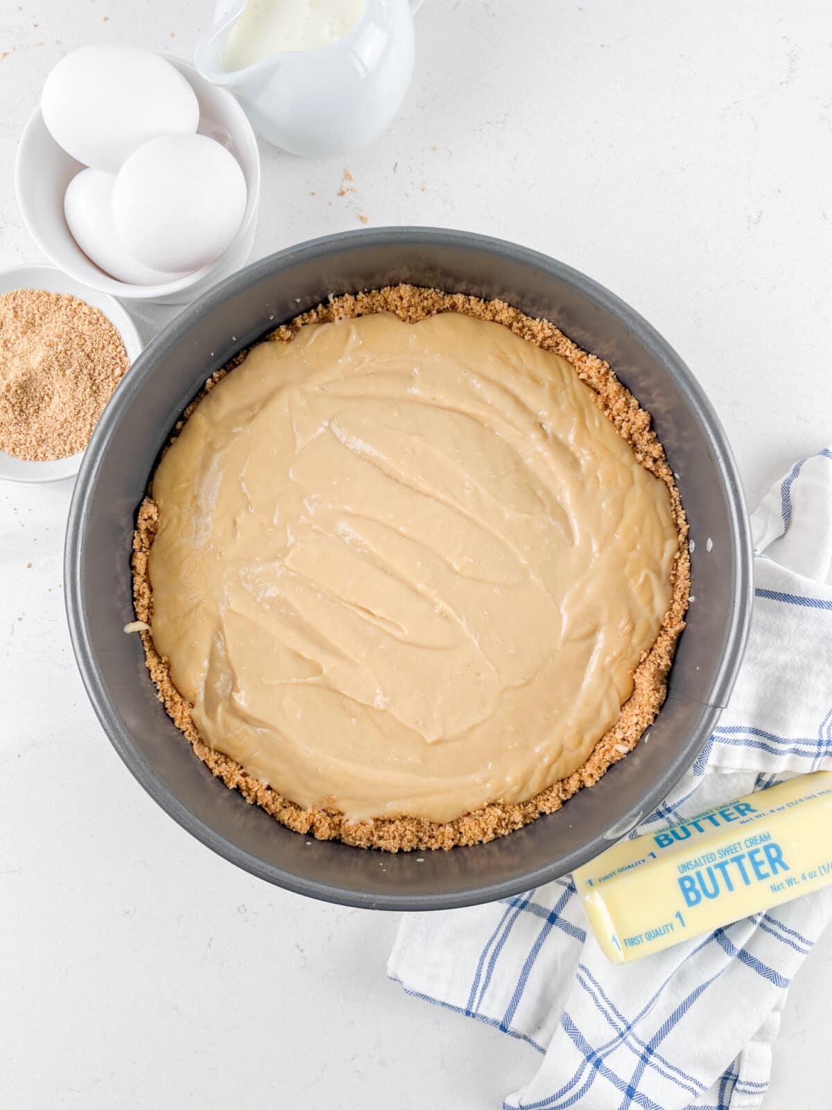 A pie crust filled with a creamy mixture sits in a round baking pan. Nearby are three eggs, a bowl of brown sugar, a stick of butter, and a blue-striped cloth. A small pitcher of milk is in the background.