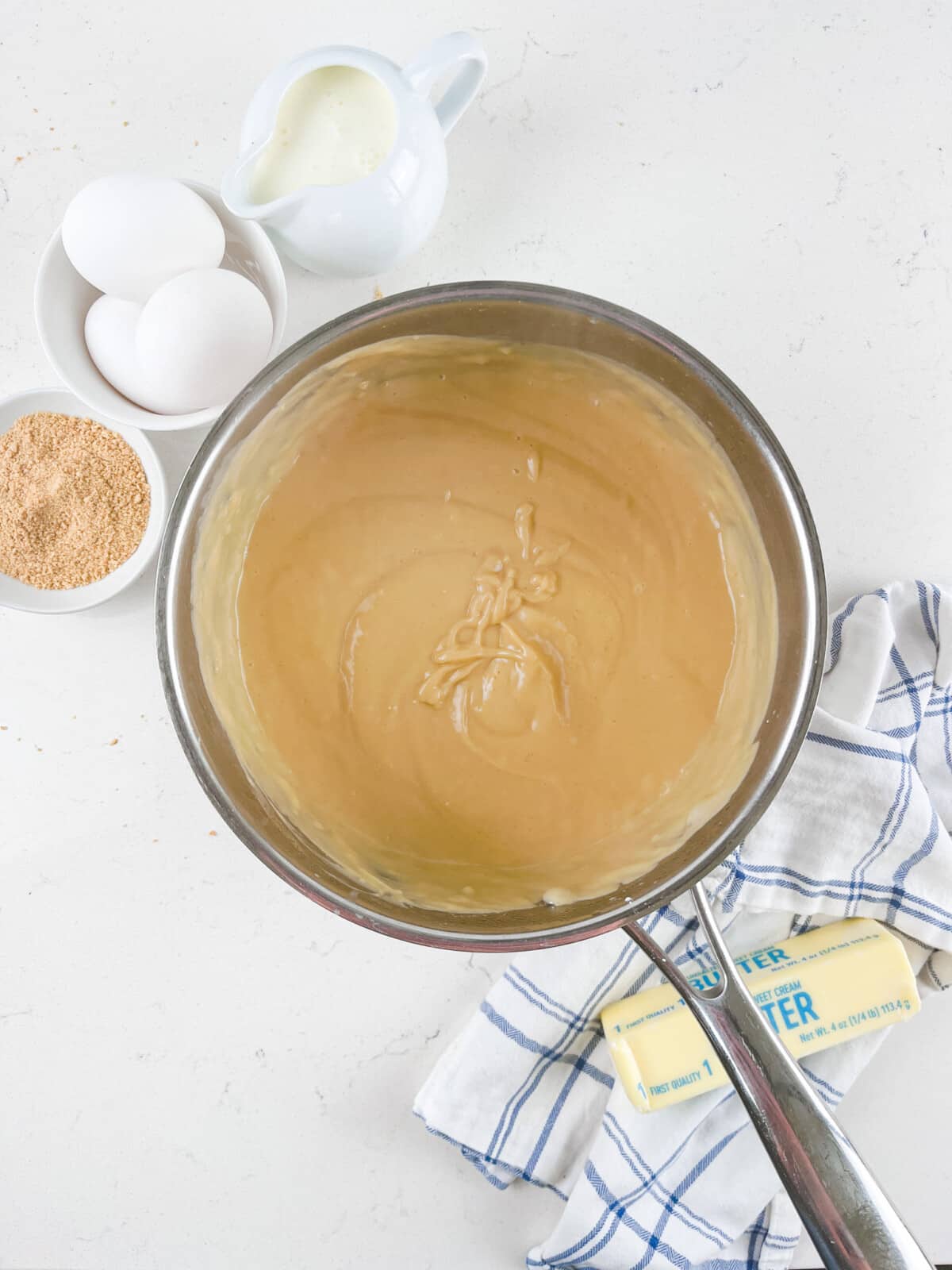 A bowl of creamy caramel sauce sits on a white surface with a blue-striped cloth beneath. Nearby are eggs, a bowl of brown sugar, a stick of butter, and a small pitcher of cream, indicating ingredients for a recipe.