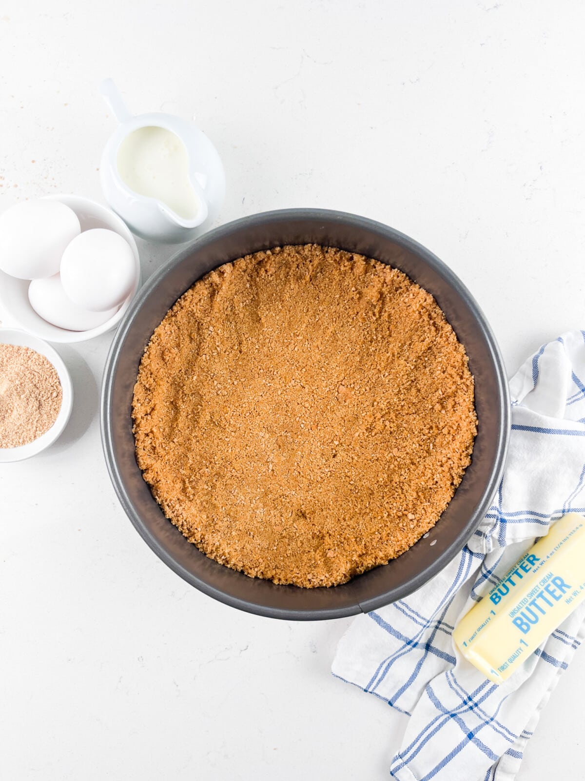 A round baking pan with a graham cracker crust is surrounded by a small pitcher of milk, three white eggs in a bowl, a bowl of brown sugar, and a wrapped stick of butter on a white and blue checked cloth, all on a white surface.