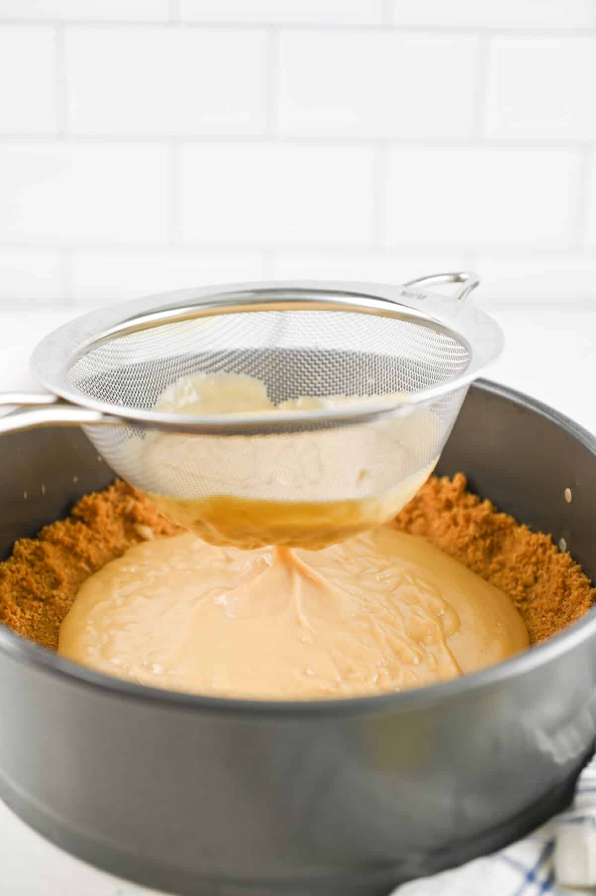 A sieve is being used to pour a smooth, light brown mixture into a springform pan with a graham cracker crust. The background features a tile wall, and a striped kitchen towel is partially visible on the counter.