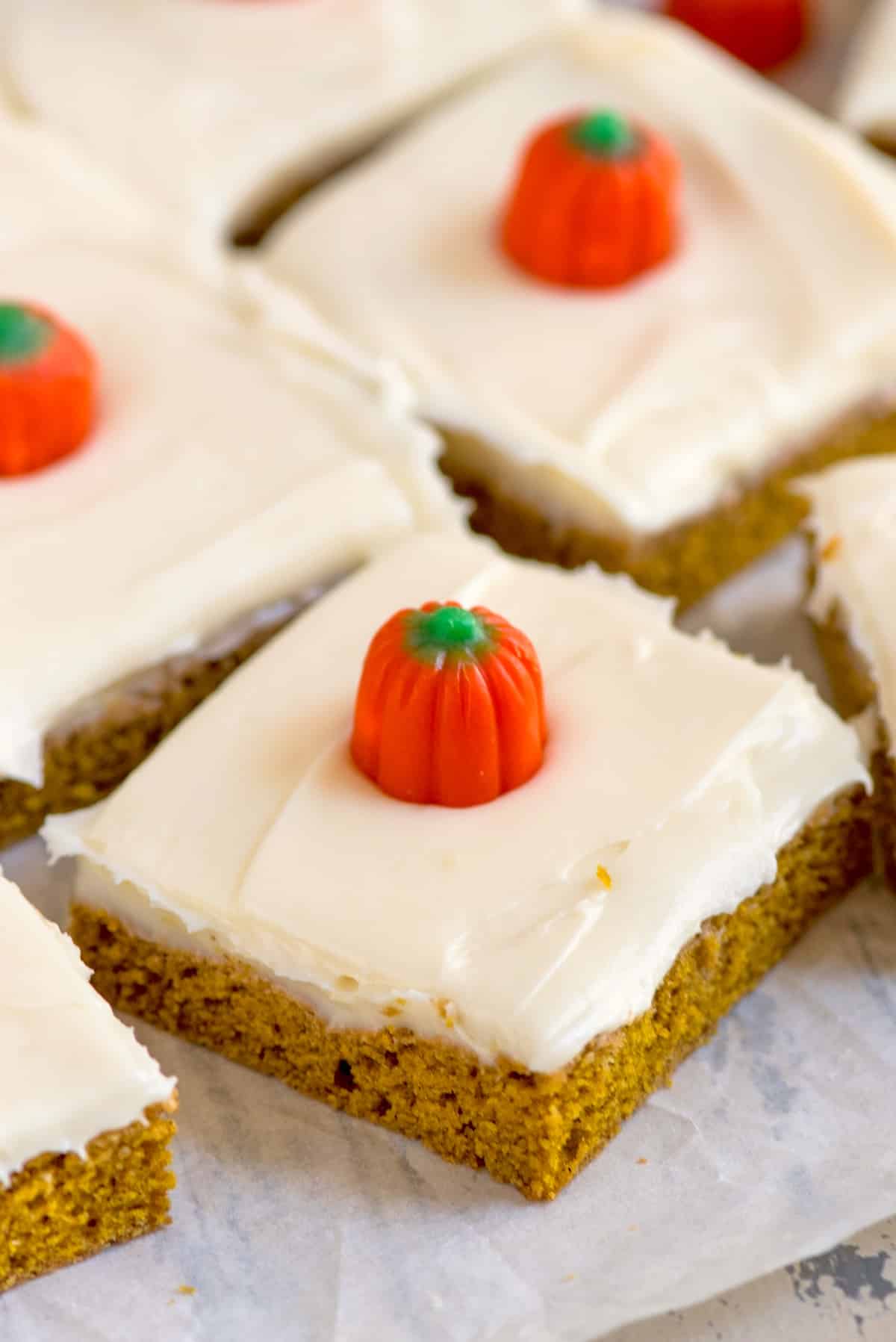 Pumpkin bars with creamy white frosting, each topped with a small candy pumpkin, are arranged on parchment paper.