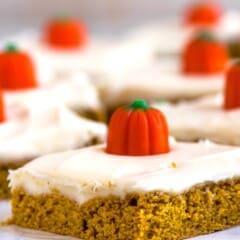 A close-up of a piece of pumpkin cake with creamy frosting and topped with a small, decorative candy pumpkin. In the background, similar pieces of cake are blurred, placed on a light wooden surface.