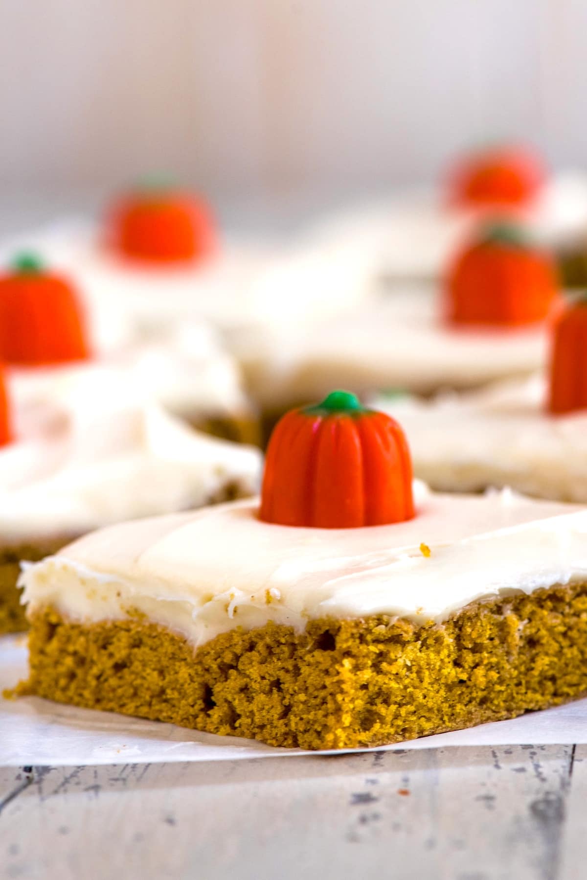 A close-up of a piece of pumpkin cake with creamy frosting and topped with a small, decorative candy pumpkin. In the background, similar pieces of cake are blurred, placed on a light wooden surface.