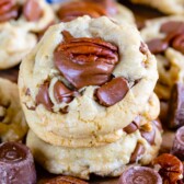 Close-up of two stacked cookies topped with chocolate chunks and pecan halves on a wooden surface, surrounded by more cookies and chocolate candies.