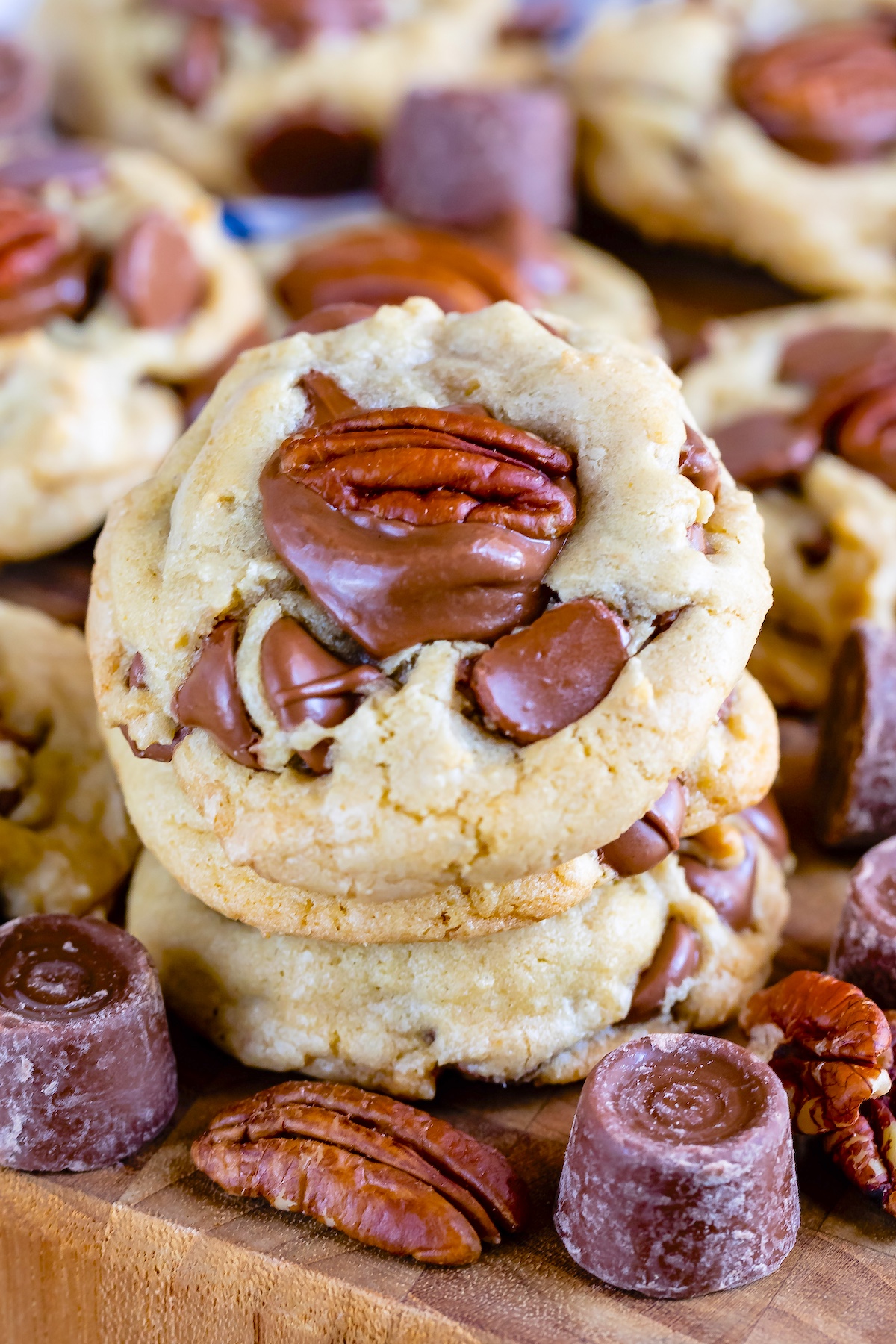 Close-up of two stacked cookies topped with chocolate chunks and pecan halves on a wooden surface, surrounded by more cookies and chocolate candies.