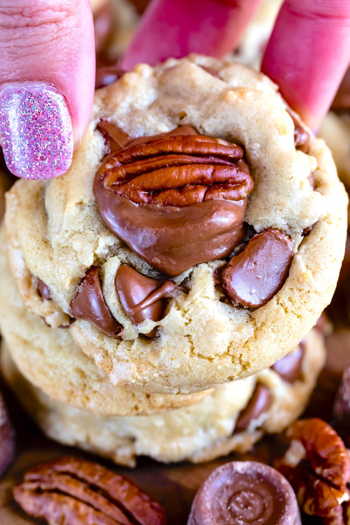 A hand with glittery pink nails holds a stack of freshly baked cookies. The cookies are topped with chocolate and pecans, showcasing a gooey, delicious texture.