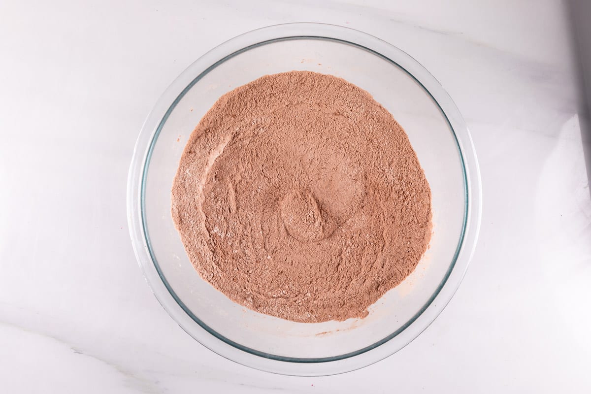 A clear glass bowl filled with a light brown, finely ground powder, likely chocolate cake mix, set on a white marble countertop. The mix appears smoothed out in the center with a slight indentation.