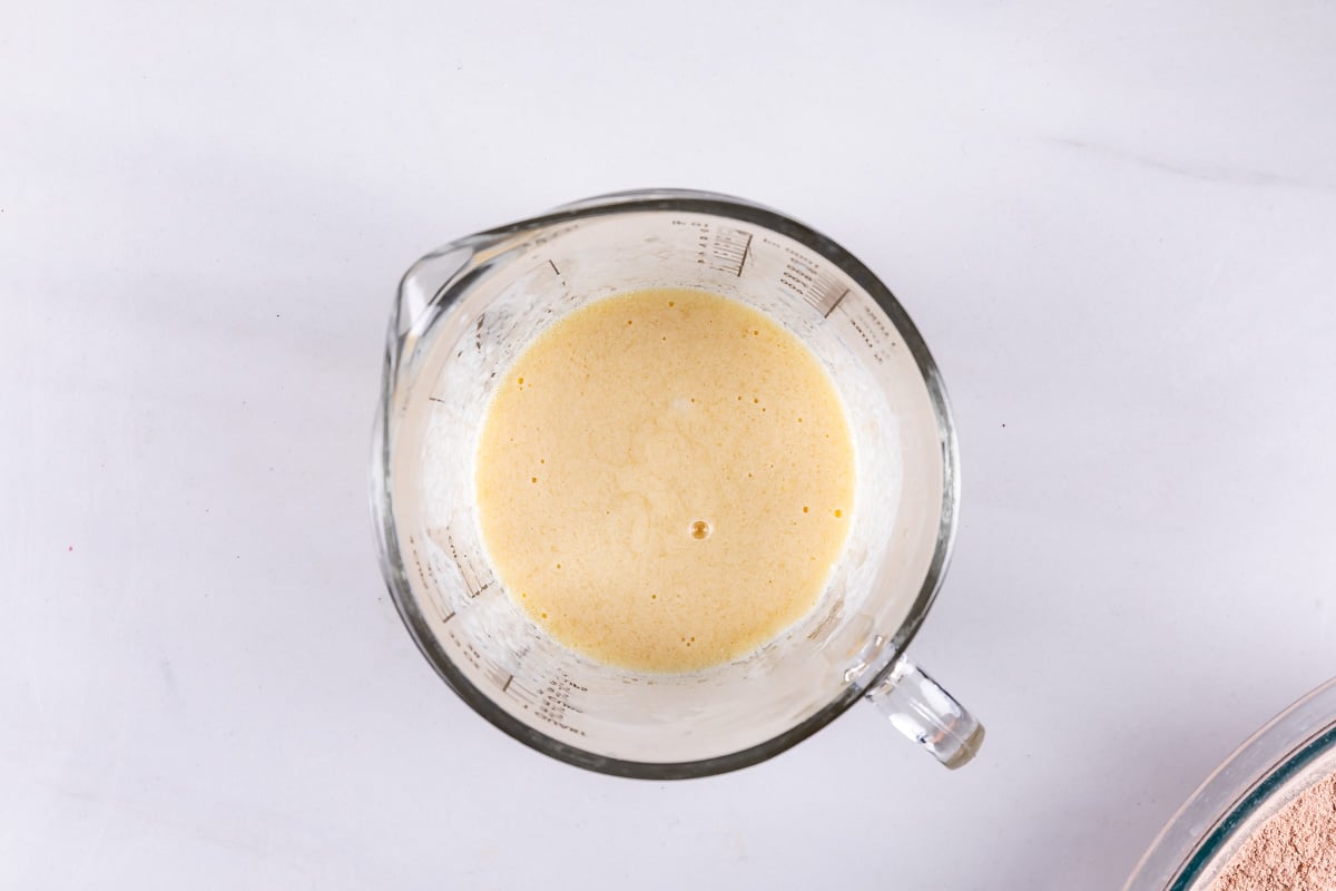 A transparent measuring cup filled with a frothy yellow batter sits on a white surface. The batter has small bubbles on its surface, indicating its well-mixed. The cup has measurement markings on its side.