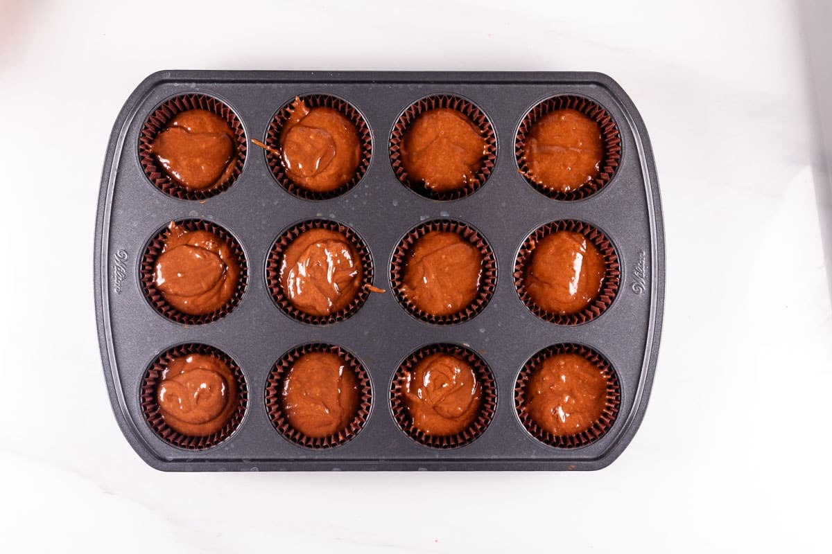 A muffin tin with twelve compartments filled with chocolate batter, ready to be baked. The tin is placed on a white countertop.