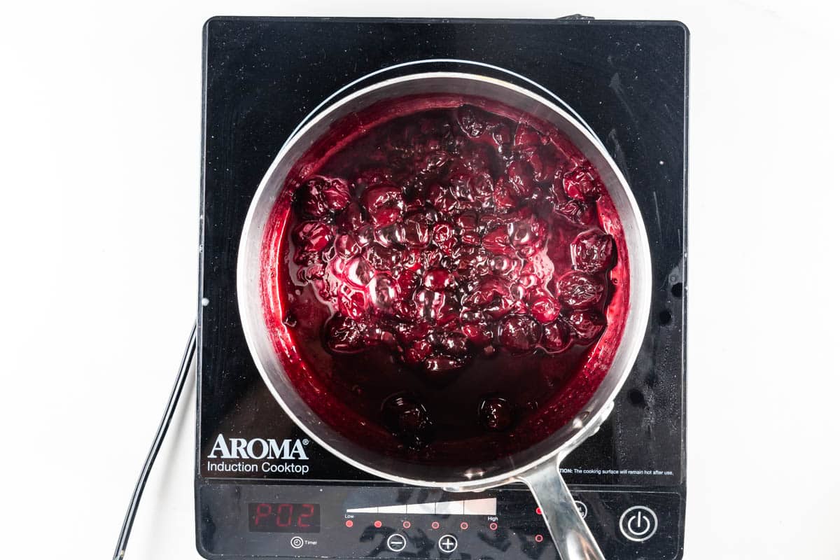 A pot of dark red cherries cooking on an Aroma induction cooktop. The cherries appear to be simmering, with bubbles visible on the surface of the liquid. The cooktop display shows 102.