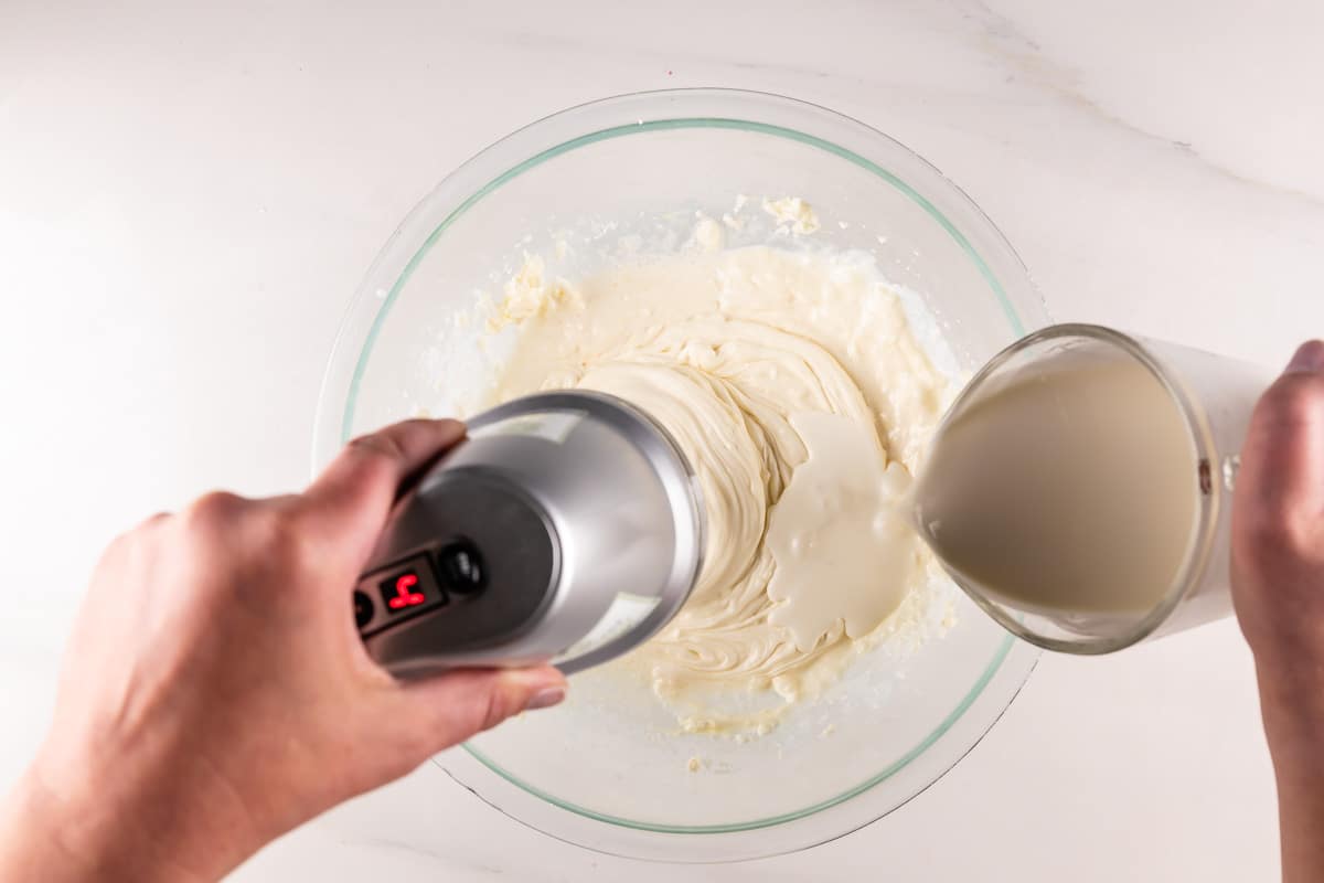 A hand mixer is being used to blend ingredients in a glass bowl, while a hand pours milk from a pitcher into the mixture. The bowl contains a creamy, pale mixture on a light countertop.