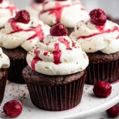 Chocolate cupcakes topped with swirls of white frosting, a red cherry on top, and drizzles of cherry sauce are arranged on a white cake stand. Small chocolate shavings and cherries are scattered around for decoration.