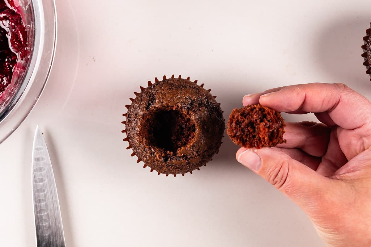 A person holds a small piece of chocolate cupcake removed from the center of a larger cupcake. A knife and a bowl with red filling are nearby on a white surface.