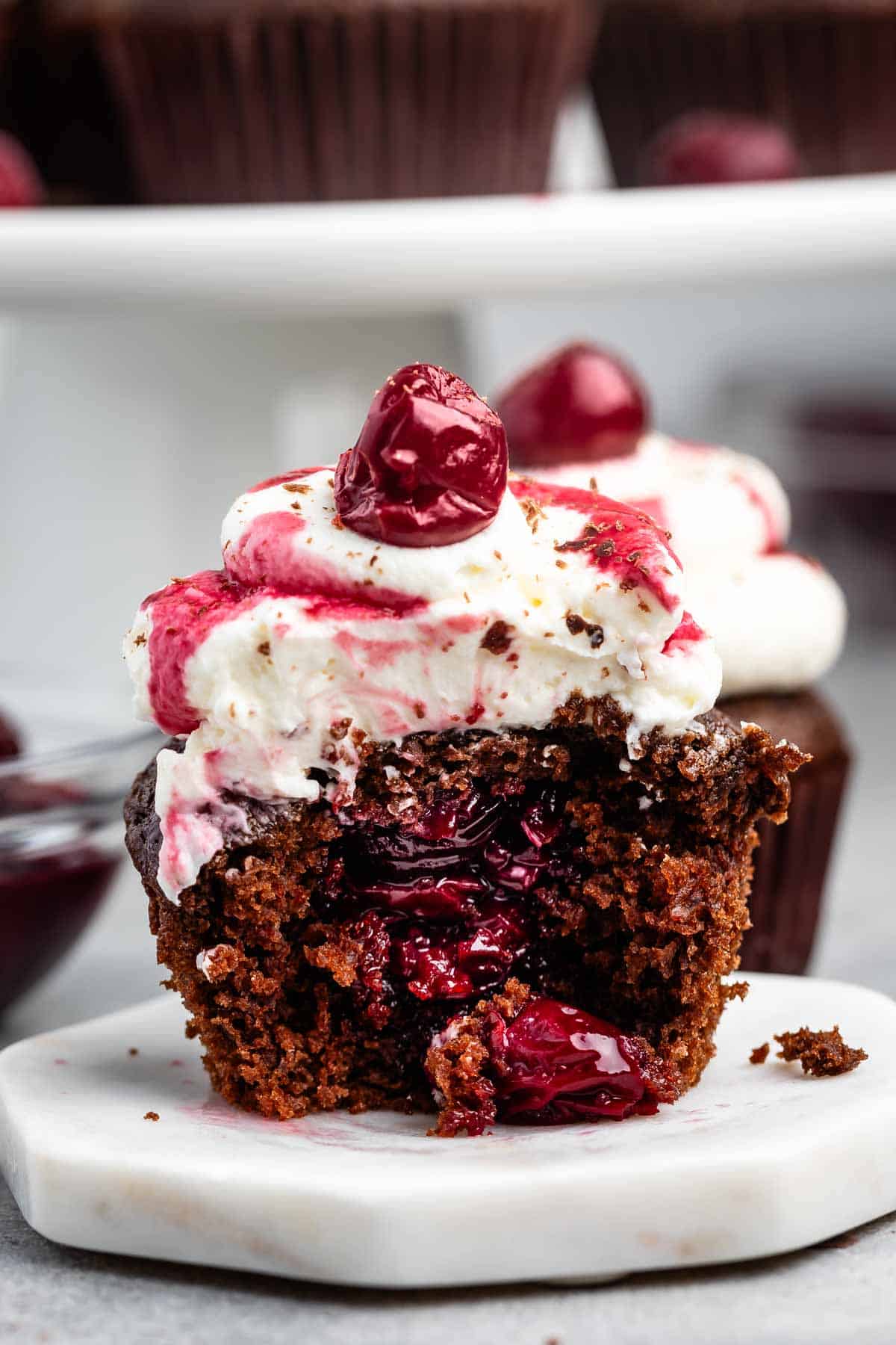 A close-up of a chocolate cupcake with a cherry filling, topped with white frosting, cherry drizzle, and a whole cherry. One bite has been taken, revealing the rich interior, and the cupcake sits on a small white plate.