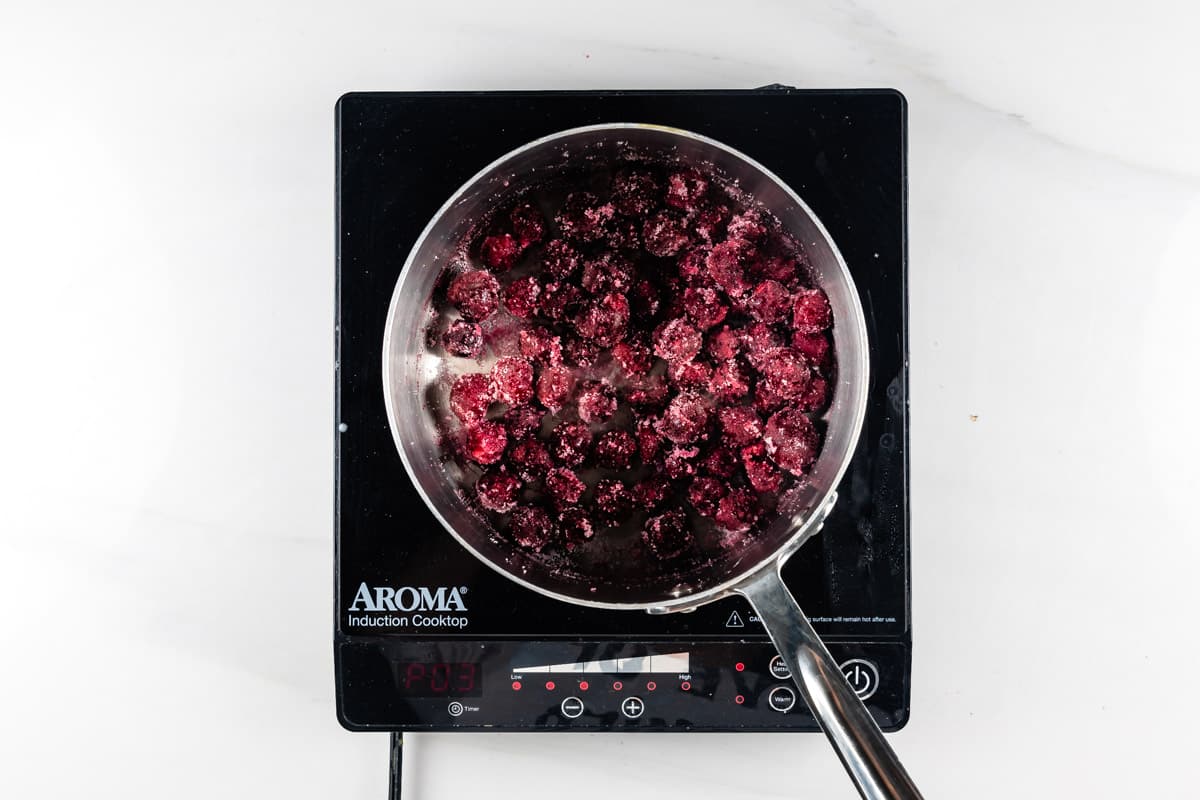 A saucepan filled with red berries is placed on an Aroma induction cooktop. The berries appear to be simmering, possibly for a sauce or jam. The cooktops digital display shows 00:3 in red. The background is a white surface.