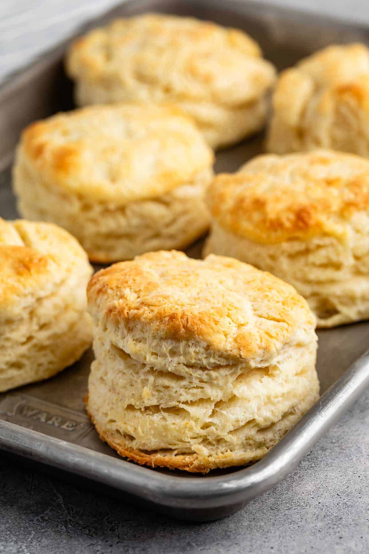 A baking tray with freshly baked golden-brown biscuits, showcasing their flaky, layered texture. The biscuits are arranged closely together on a silver tray, on a gray surface.