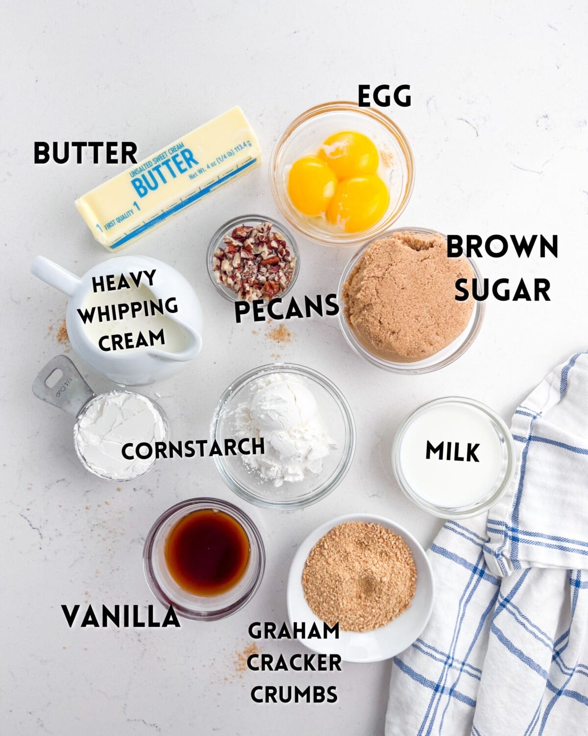 Ingredients laid out on a white surface include a stick of butter, heavy whipping cream, chopped pecans, cornstarch, vanilla, egg yolks, brown sugar, milk, and graham cracker crumbs, with a blue and white checkered cloth nearby.