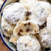 A bowl filled with powdered sugar-coated cookie dough balls, some with chocolate chips visible. One dough ball is halved, revealing a soft, chocolate chip-studded interior. The bowl has a colorful rim.