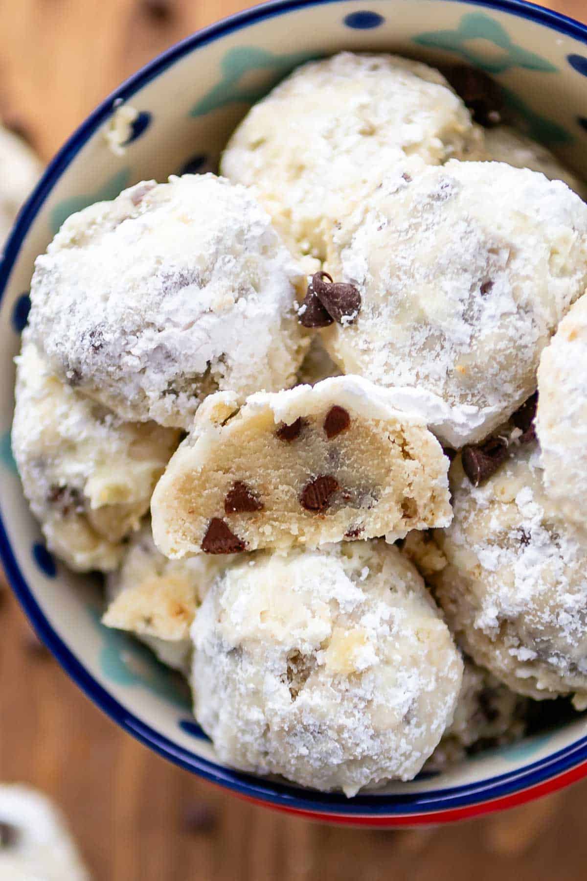 A bowl filled with powdered sugar-coated cookie dough balls, some with chocolate chips visible. One dough ball is halved, revealing a soft, chocolate chip-studded interior. The bowl has a colorful rim.