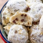 A bowl filled with powdered sugar-covered cookies, some of which have chocolate chips visible. One cookie is broken in half to reveal the chocolate chips inside. The bowl has a colorful floral pattern.