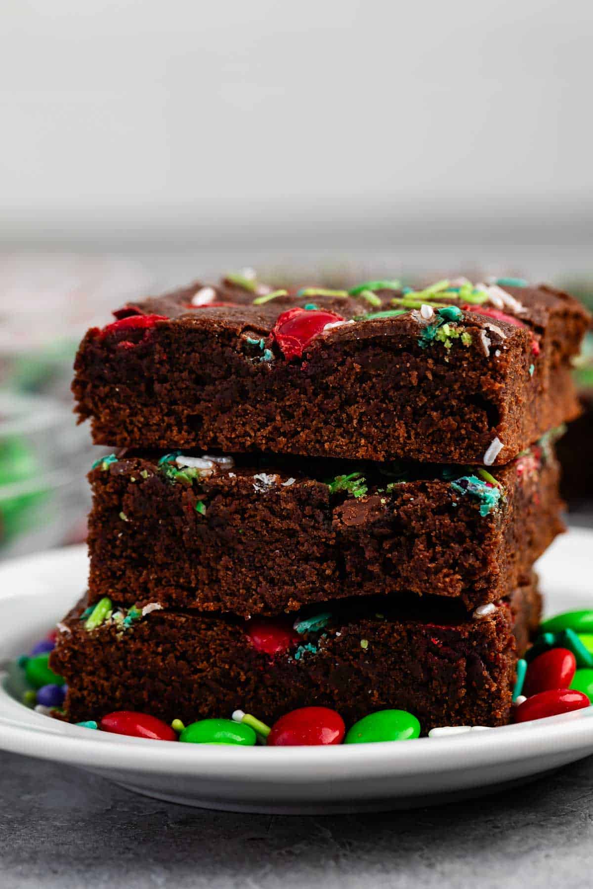 Three stacked chocolate brownies with colorful candy pieces on top, placed on a white plate adorned with red and green candies. The background is blurred, highlighting the rich texture of the brownies.