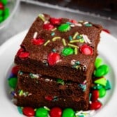 A stack of three chocolate brownies topped with colorful candy-coated chocolates and sprinkles sits on a white plate. More candies are scattered around the plate, with additional brownies visible in the background.