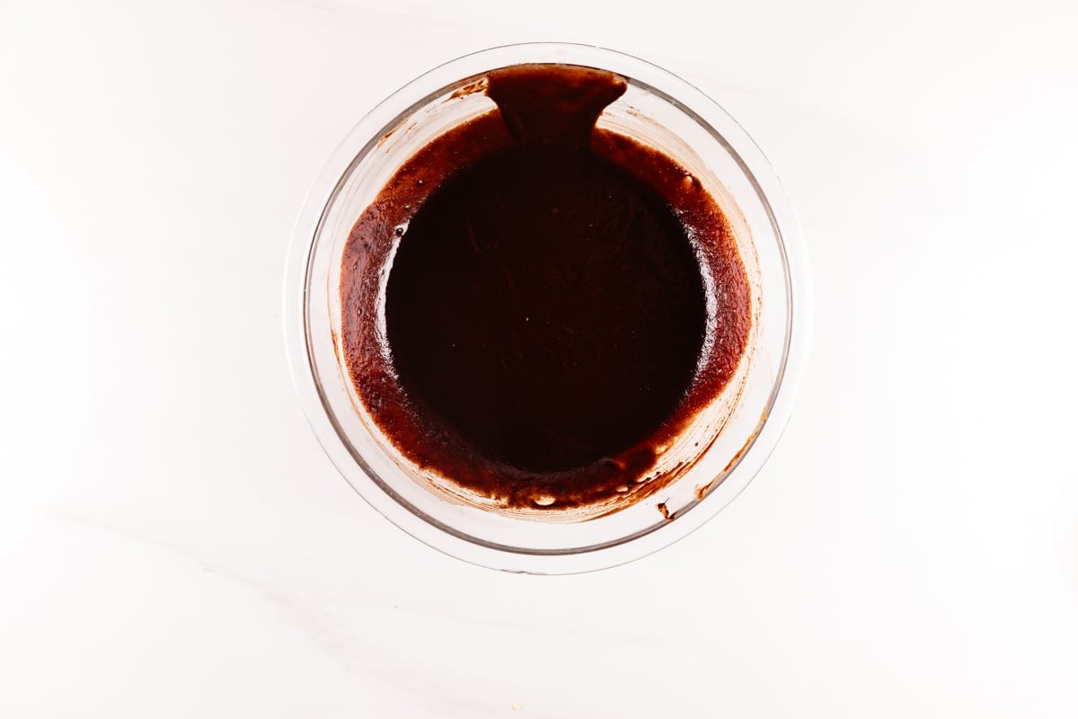 A clear glass bowl filled with smooth, rich chocolate batter on a white background. The batter has a glossy texture and covers the bottom of the bowl evenly.