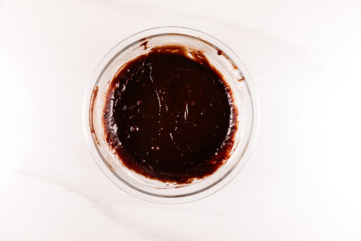 A glass bowl filled with smooth, dark chocolate cake batter sits on a marble surface. The rich, glossy mixture is evenly spread within the bowl, ready for baking.