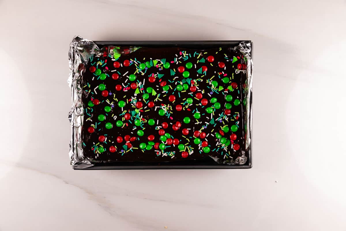 A rectangular tray of chocolate brownies covered with red and green candy-coated chocolates and colorful sprinkles, lined with aluminum foil on a white background.