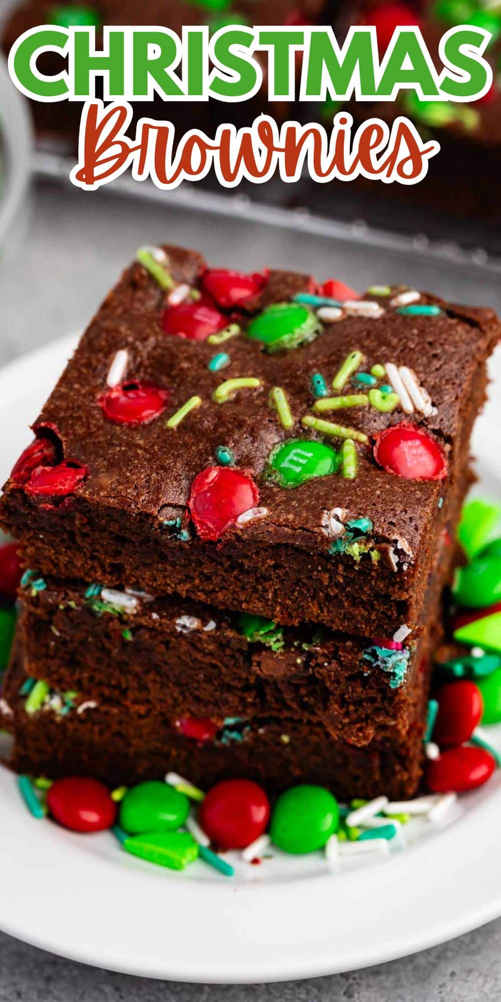 A stack of three chocolate brownies on a white plate, topped with red and green candies and colorful sprinkles. The image has Christmas Brownies written at the top.