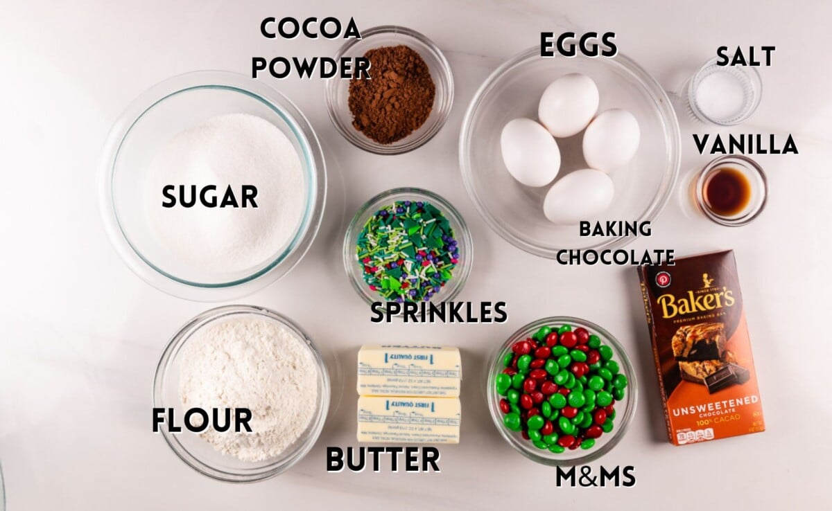 A baking setup displays ingredients: bowls of sugar, cocoa powder, and flour, four eggs, a butter stick, a bottle of vanilla, a pinch of salt, a bar of unsweetened baking chocolate, colorful sprinkles, and red and green M&Ms.