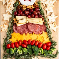 A festive charcuterie board arranged in the shape of a Christmas tree, featuring a brie with a star-shaped jelly, cheese slices, salami, grape tomatoes, broccoli, grapes, and rosemary sprigs. Surrounded by star-shaped crackers.