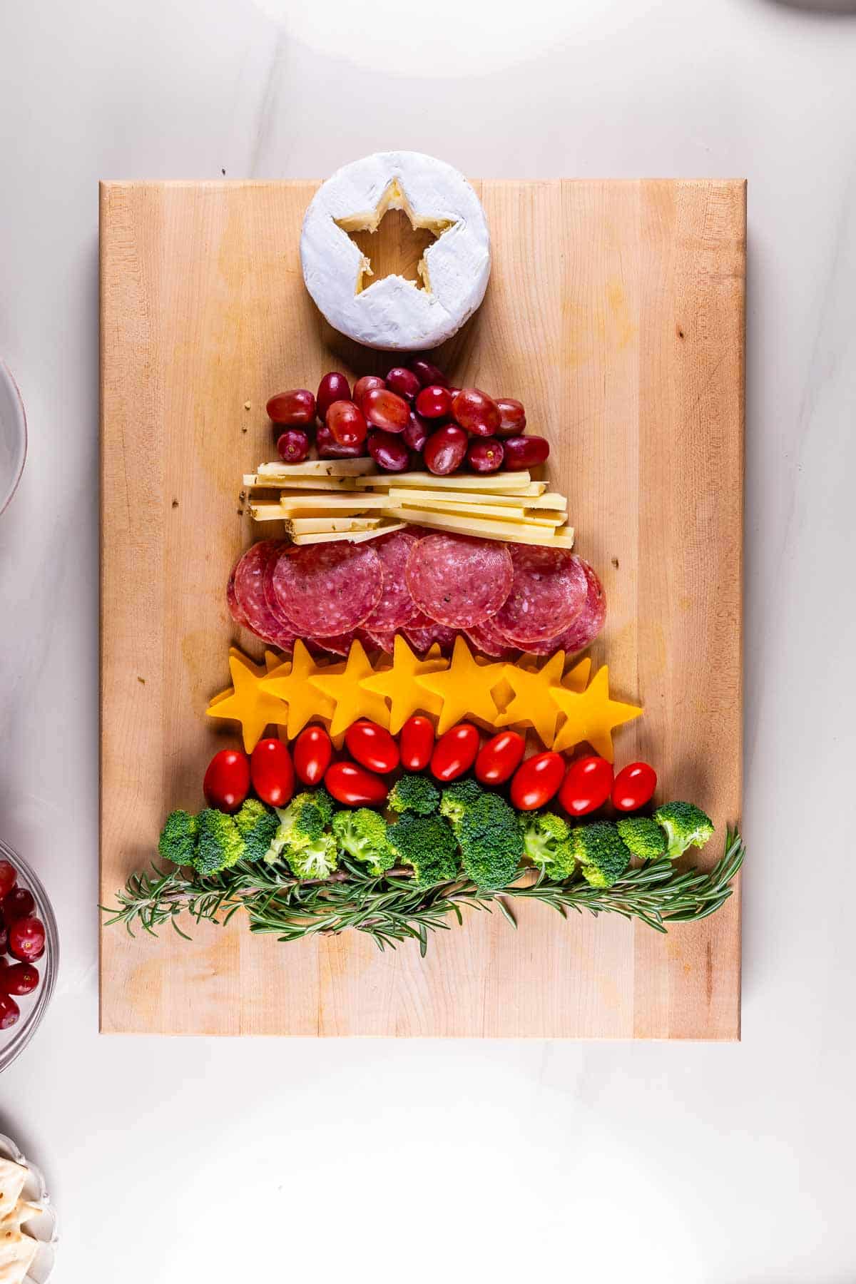 Charcuterie board arranged in the shape of a Christmas tree. From top to bottom: a round of cheese with a star cutout, grapes, salami, cheese slices, cherry tomatoes, broccoli, and rosemary sprigs on a wooden board.