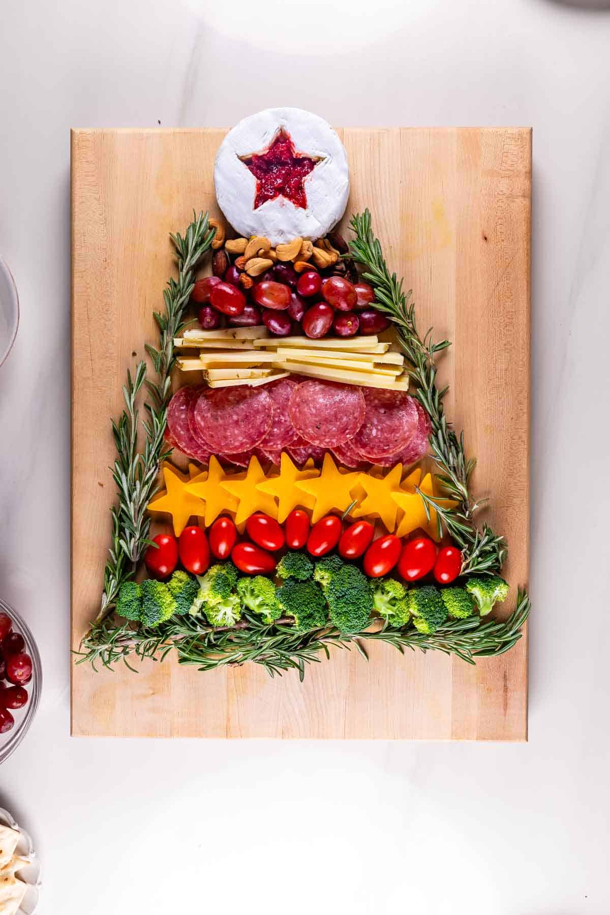 Charcuterie board arranged in a Christmas tree shape with a star-shaped cheese on top. Layers include cheese slices, cherry tomatoes, broccoli, meats, nuts, and herbs on a wooden board.