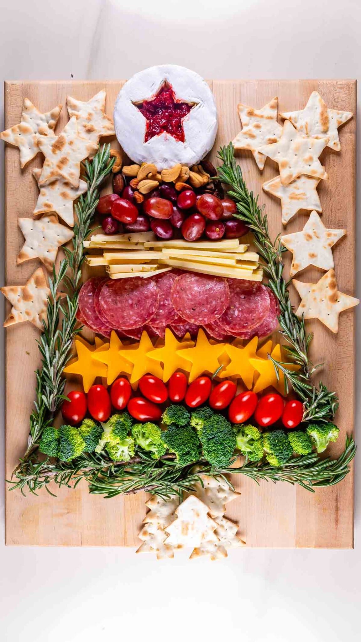 Festive charcuterie board arranged like a Christmas tree with a brie and jam top, crackers shaped as stars, nuts, grapes, cheese, salami, broccoli, tomatoes, and rosemary sprigs on a wooden background.