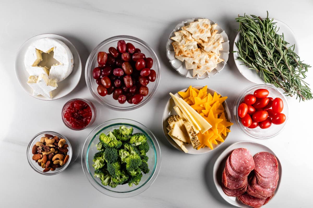 A variety of foods on a white surface, including brie cheese, grapes, crackers, rosemary, cherry tomatoes, sliced cheddar, cured meats, mixed nuts, broccoli, and a small dish of jam.