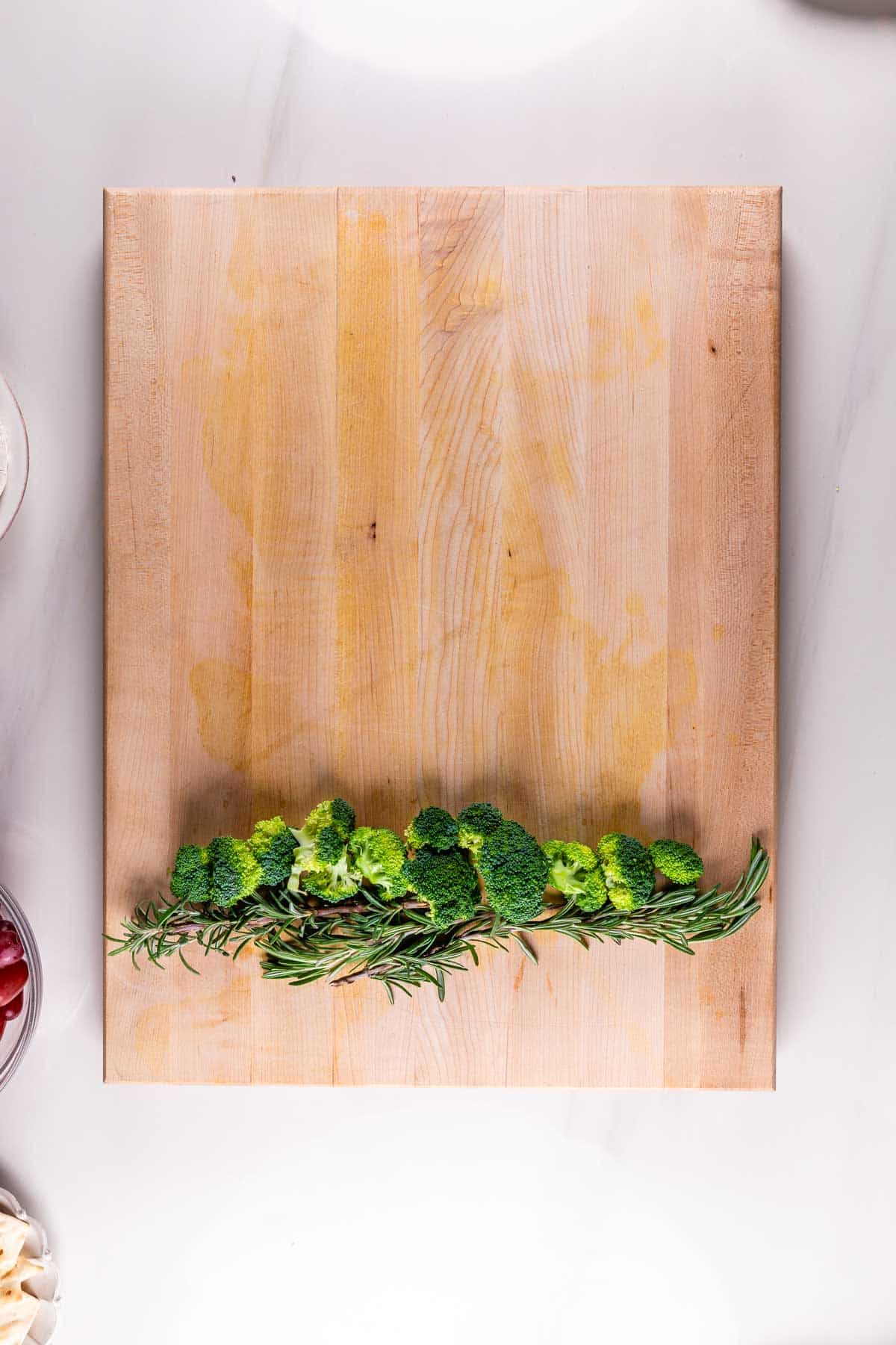 A wooden cutting board with a line of broccoli and sprigs of rosemary arranged neatly along the bottom edge. A few bowls with ingredients are partially visible around the edges.