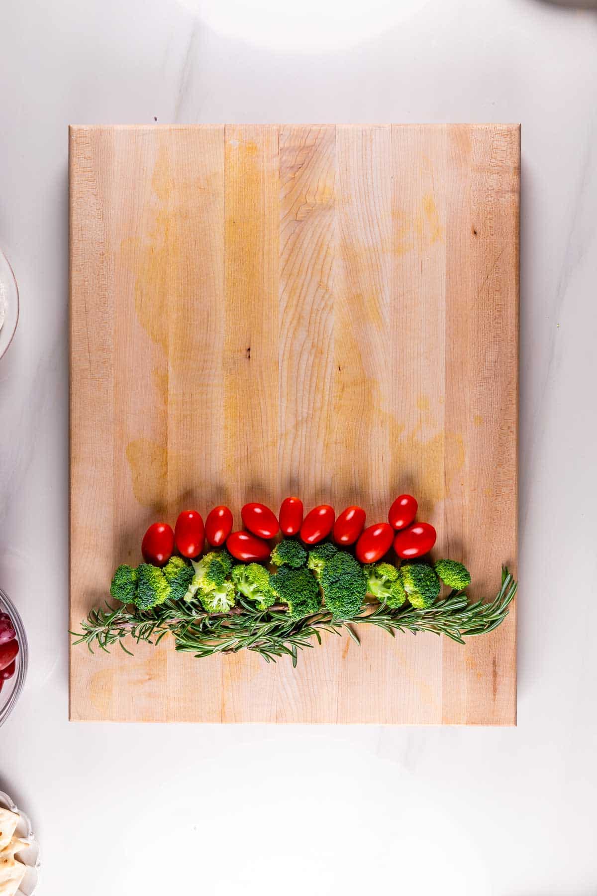 A wooden cutting board displays a neat row of fresh broccoli florets, cherry tomatoes, and sprigs of rosemary against a plain white background. Small portions of other ingredients can be seen in the corners.